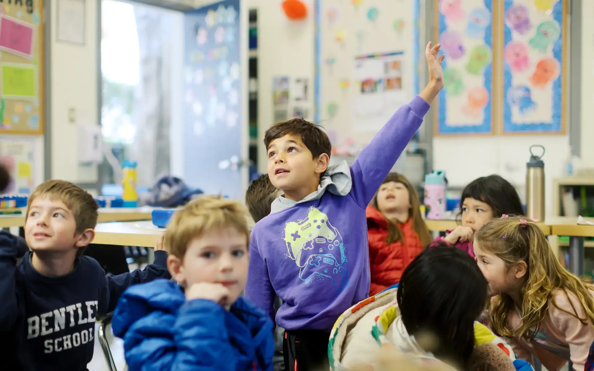 A 1st-grade boy raises his hand while sitting with classmates on a classroom rug. 