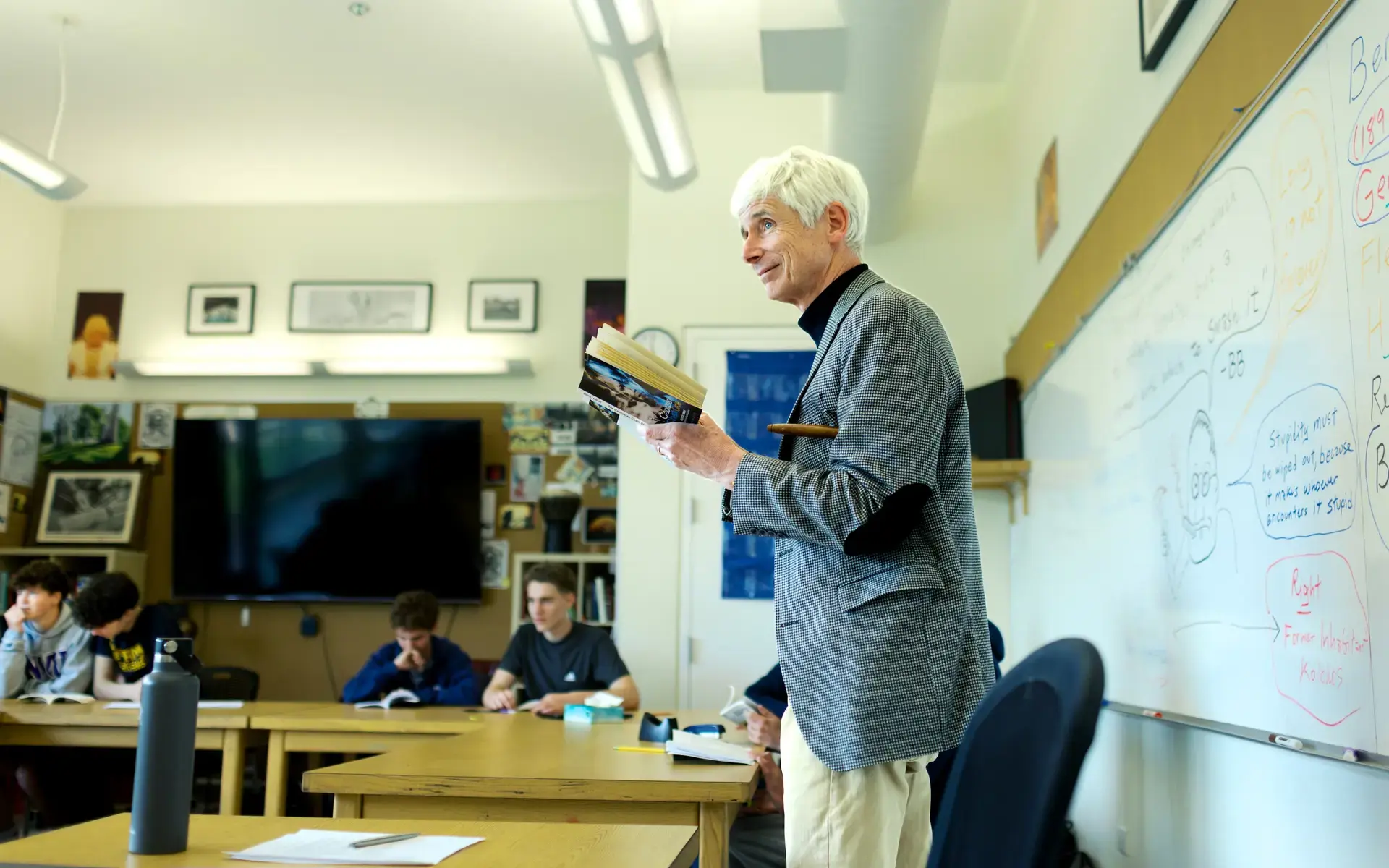 An Upper School English teacher holds a book in front of students, with a whiteboard behind him. 