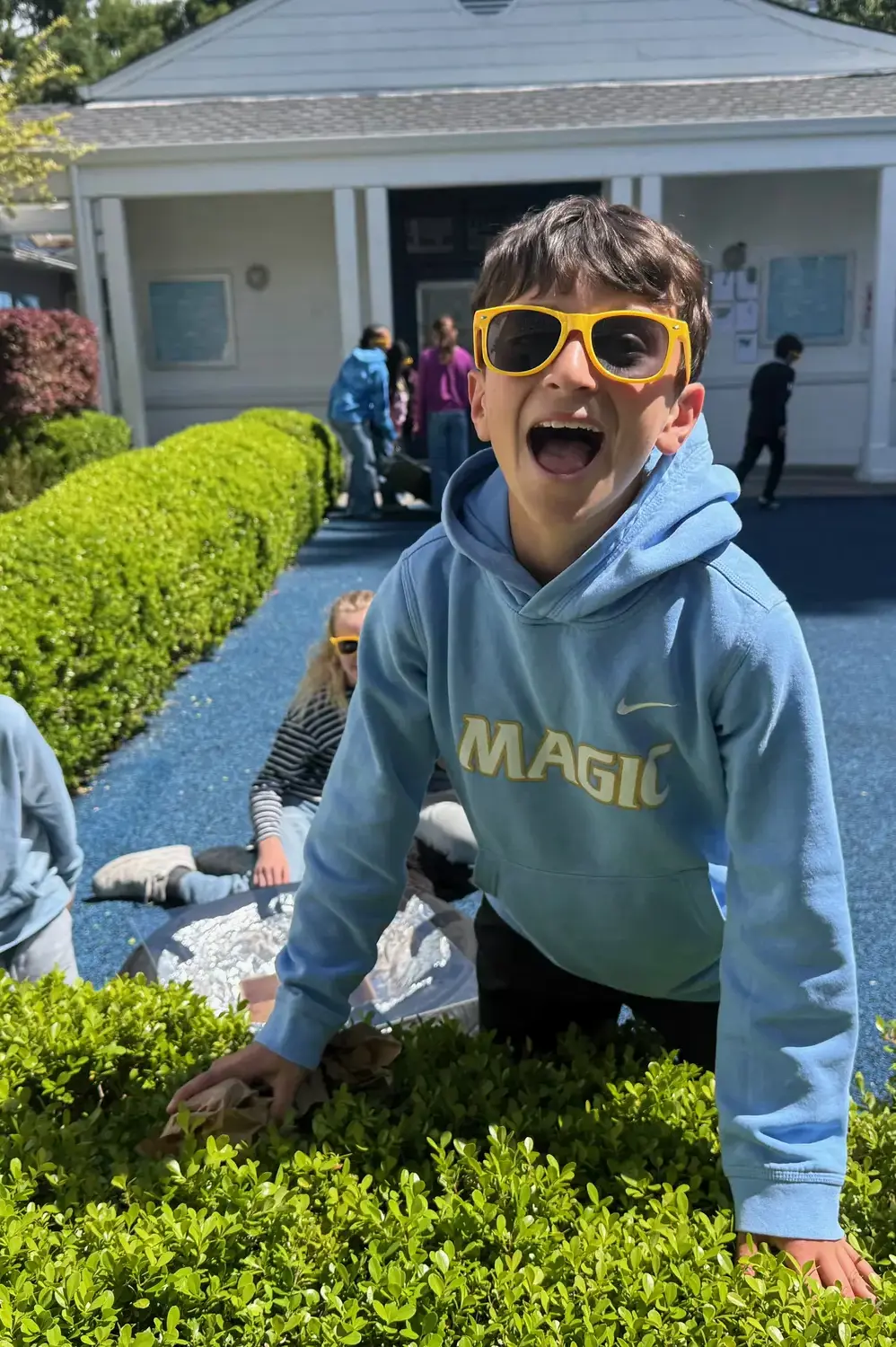 A 5th grade student smiles and laughs at the camera as he leans on the hedge in front of the Library. 