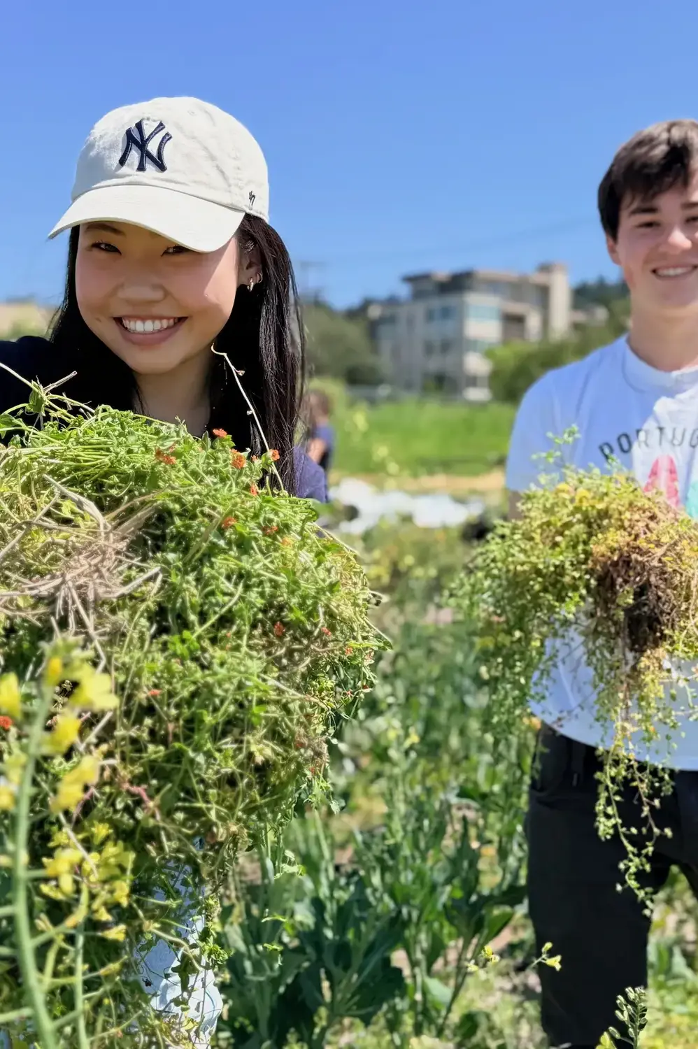 Two Upper School students hold out armfuls of weeds they've just picked from a community garden. 