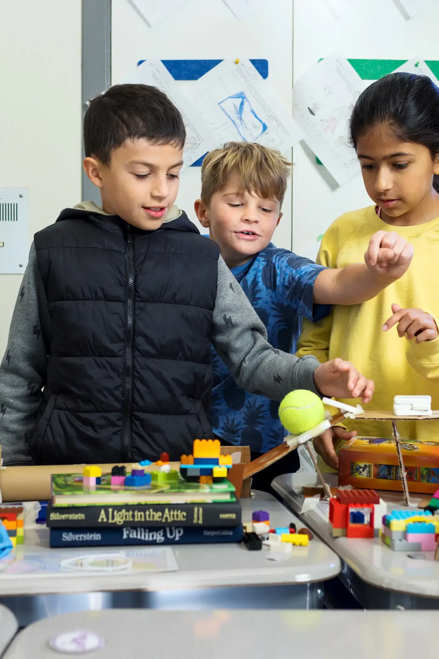 Three 3rd-graders huddle and reach out over their simple machines.