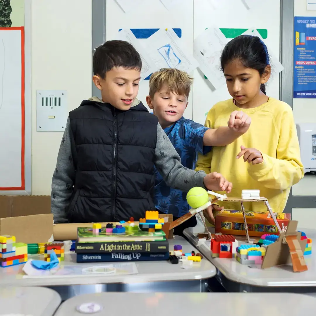 Three 3rd-graders huddle around a simple machine together in a classroom. 