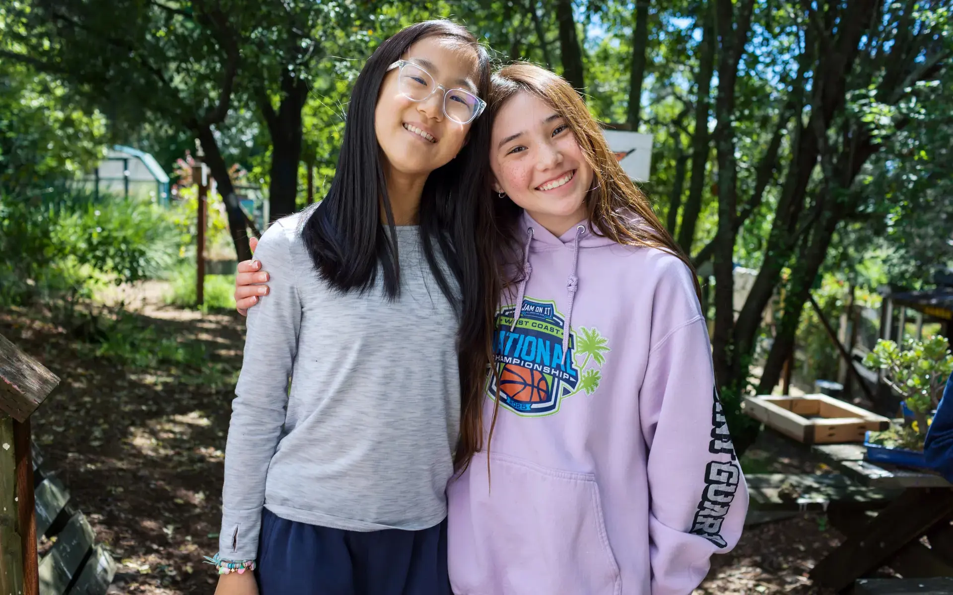 Two Middle School girls smile with their arms around one another in the Hiller Garden. 